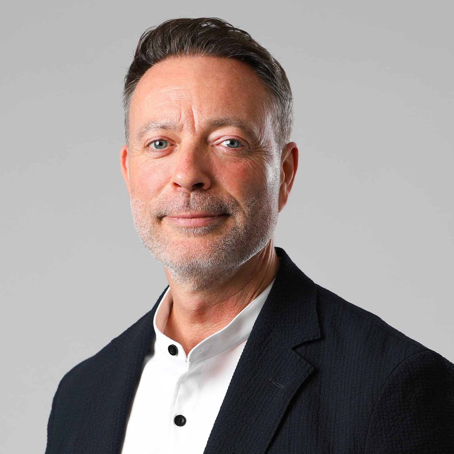 A man with short hair and a beard, wearing a black jacket over a white shirt, smiles.