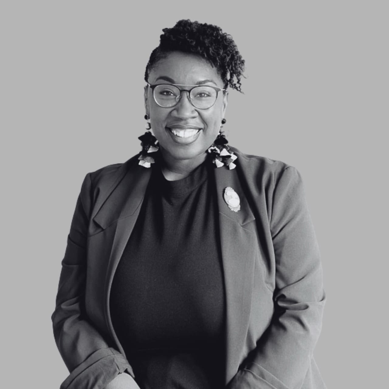 Professional woman in blazer and glasses smiling at camera, wearing statement earrings and pin