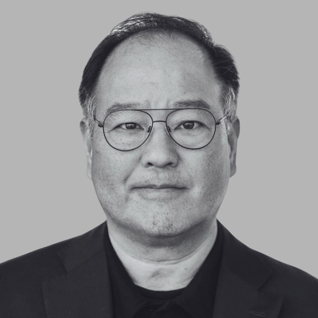 Portrait of a man wearing glasses, dressed in a black suit against a gray background.