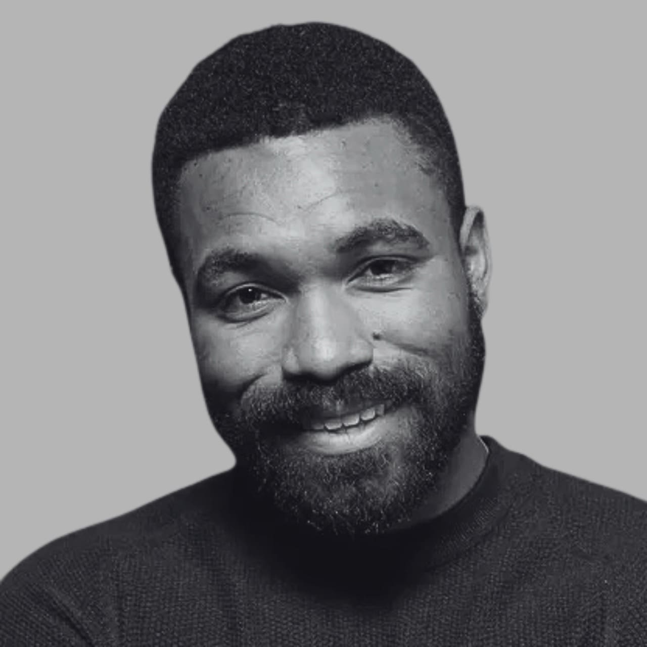 A smiling person with a beard wearing a dark sweater against a gray background.