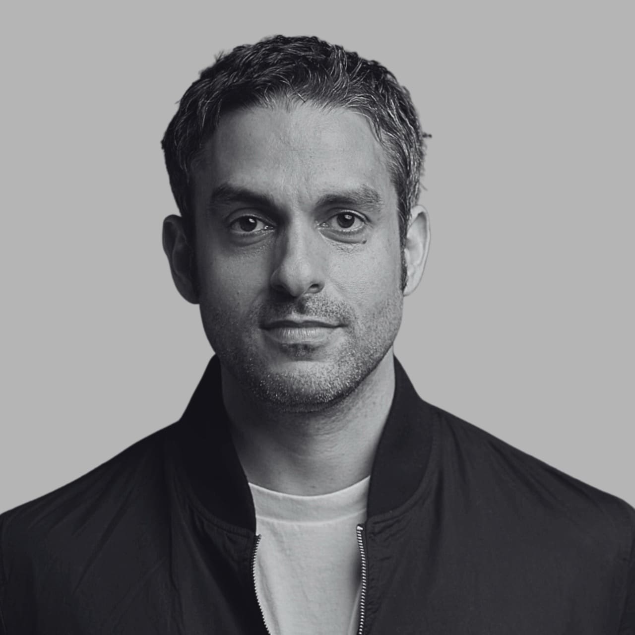 Portrait of a man with short hair, wearing a black jacket and white shirt, against a gray background.