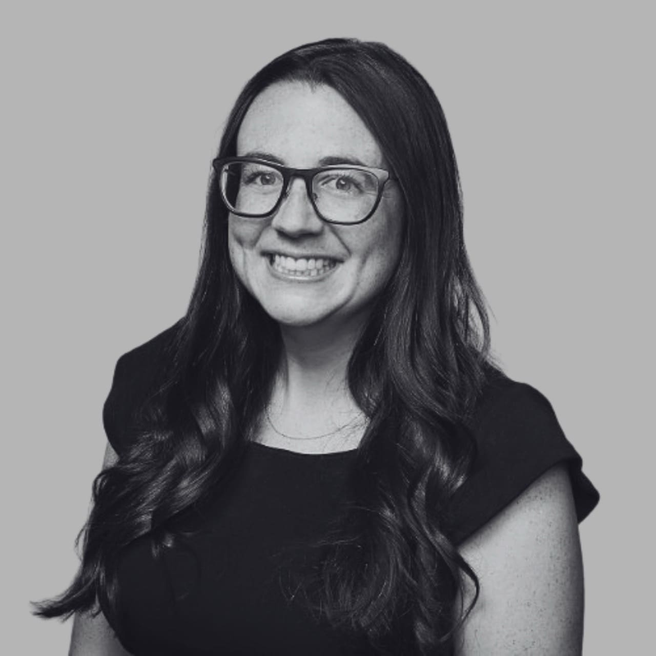 A smiling person with long hair wearing glasses and a black top against a gray background.