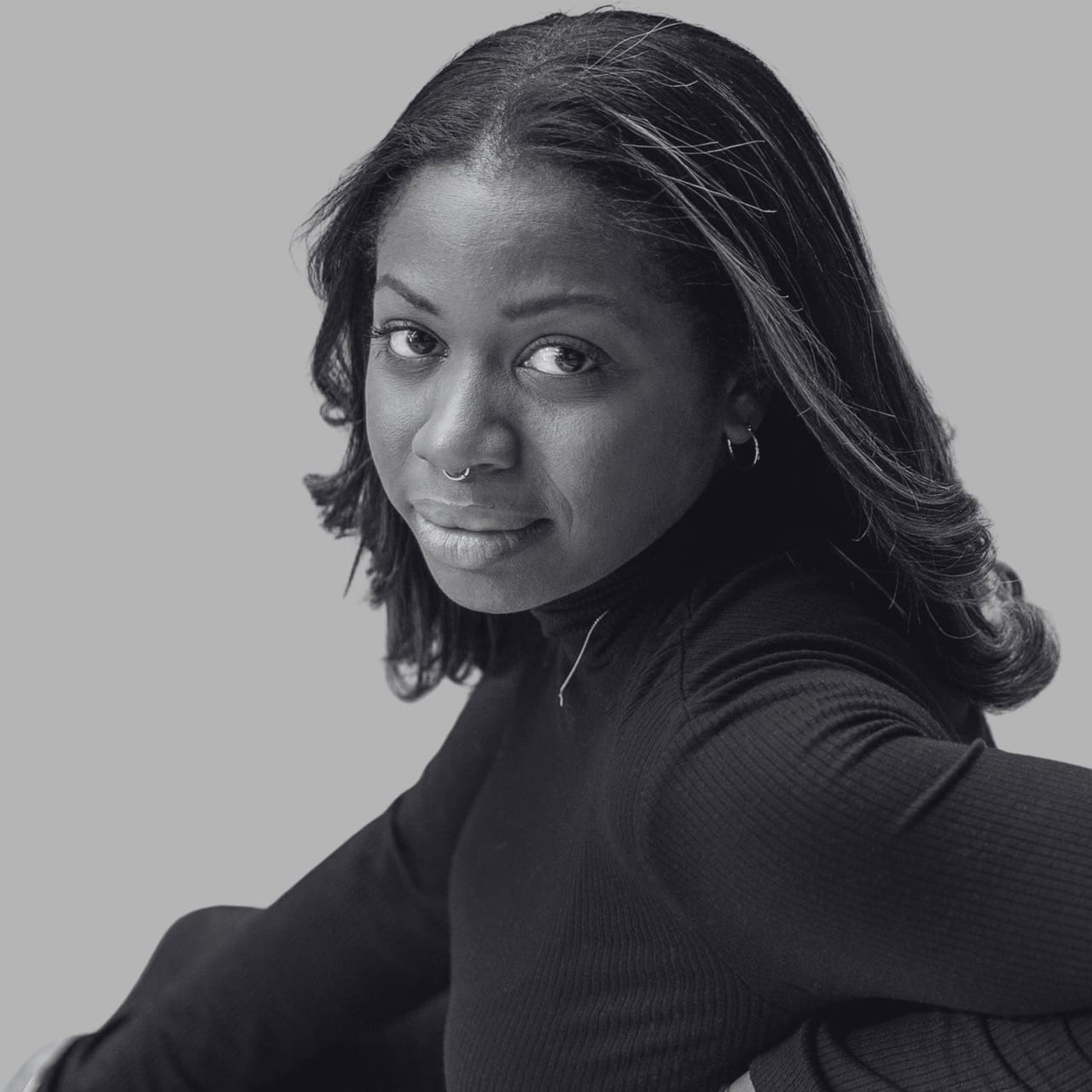 A woman with long hair and a septum piercing gazes thoughtfully at the camera.
