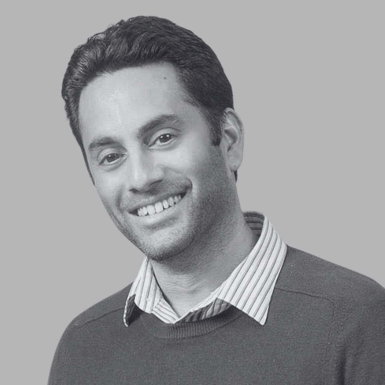 Smiling man in a grey sweater and striped shirt against a grey background.