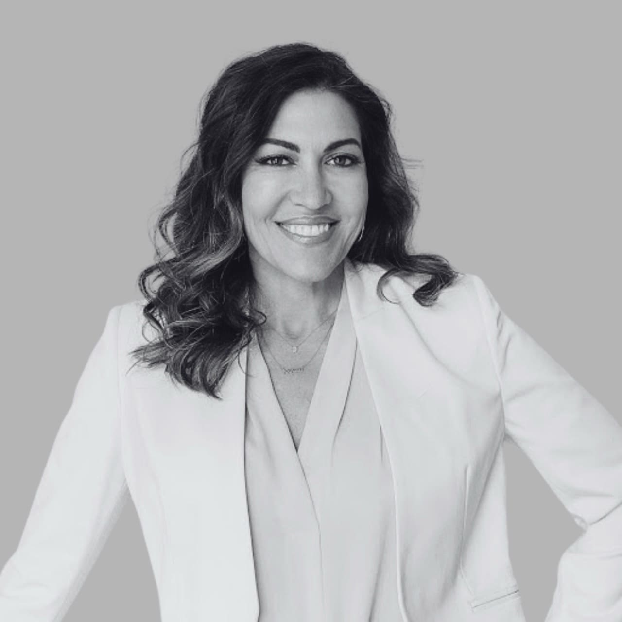 A smiling woman in a white blazer poses confidently against a gray background.