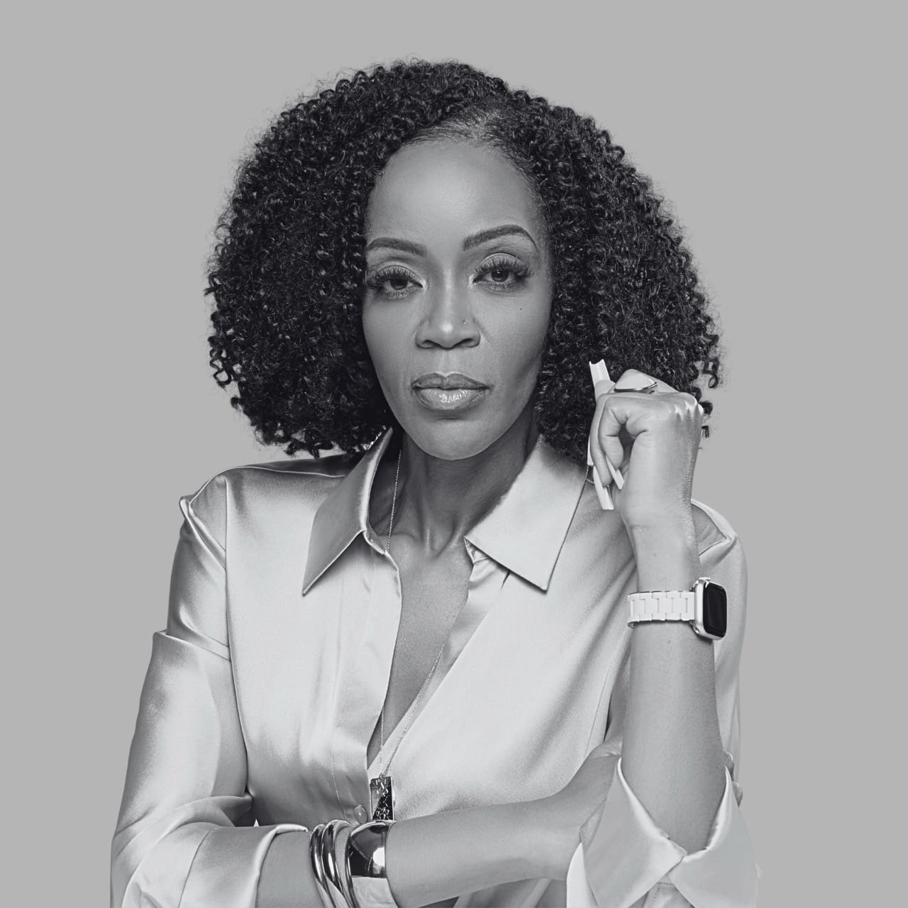 Confident woman in a silver shirt, with curly hair, holding a pen and wearing a watch.