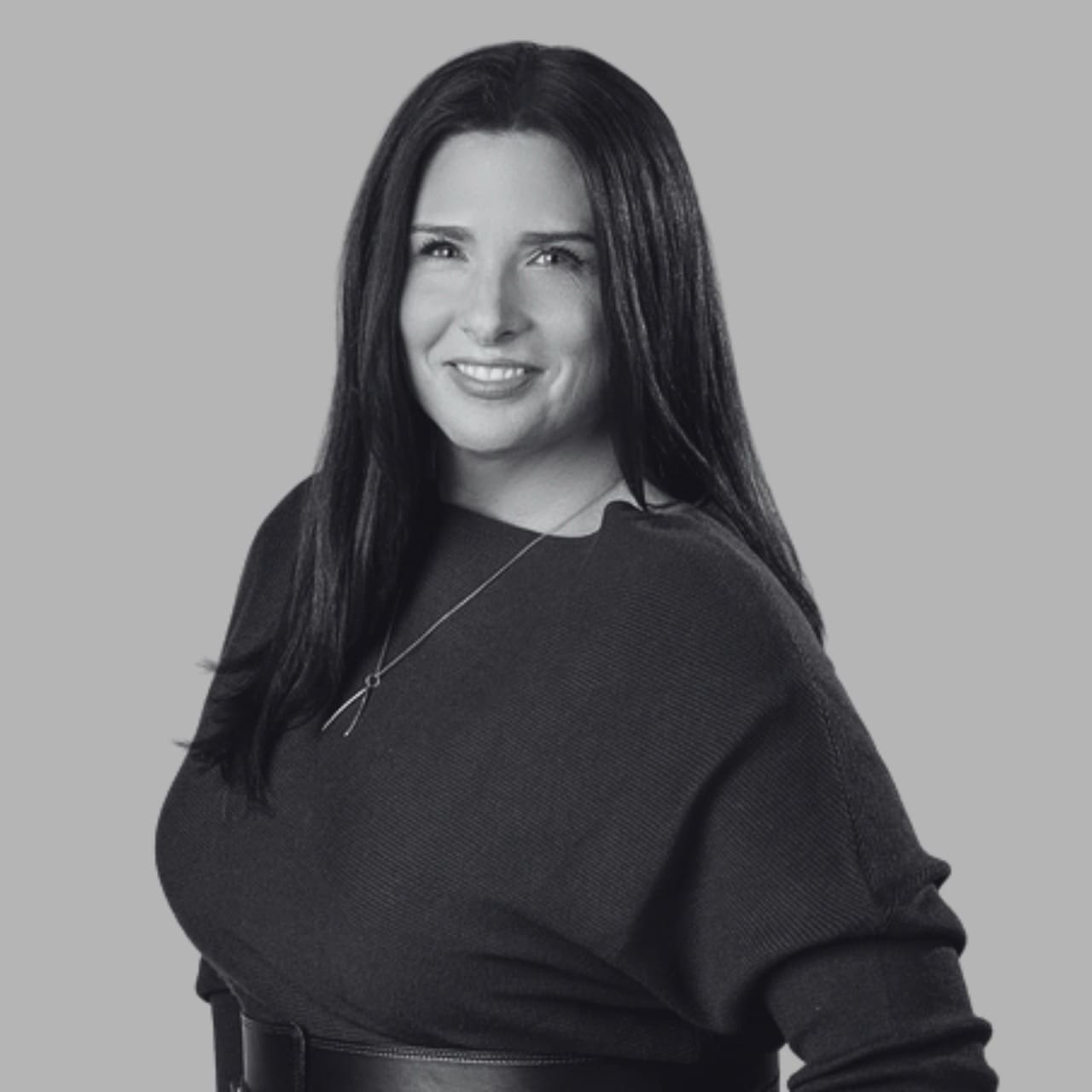 A woman with long dark hair and a black sweater smiles against a gray background.