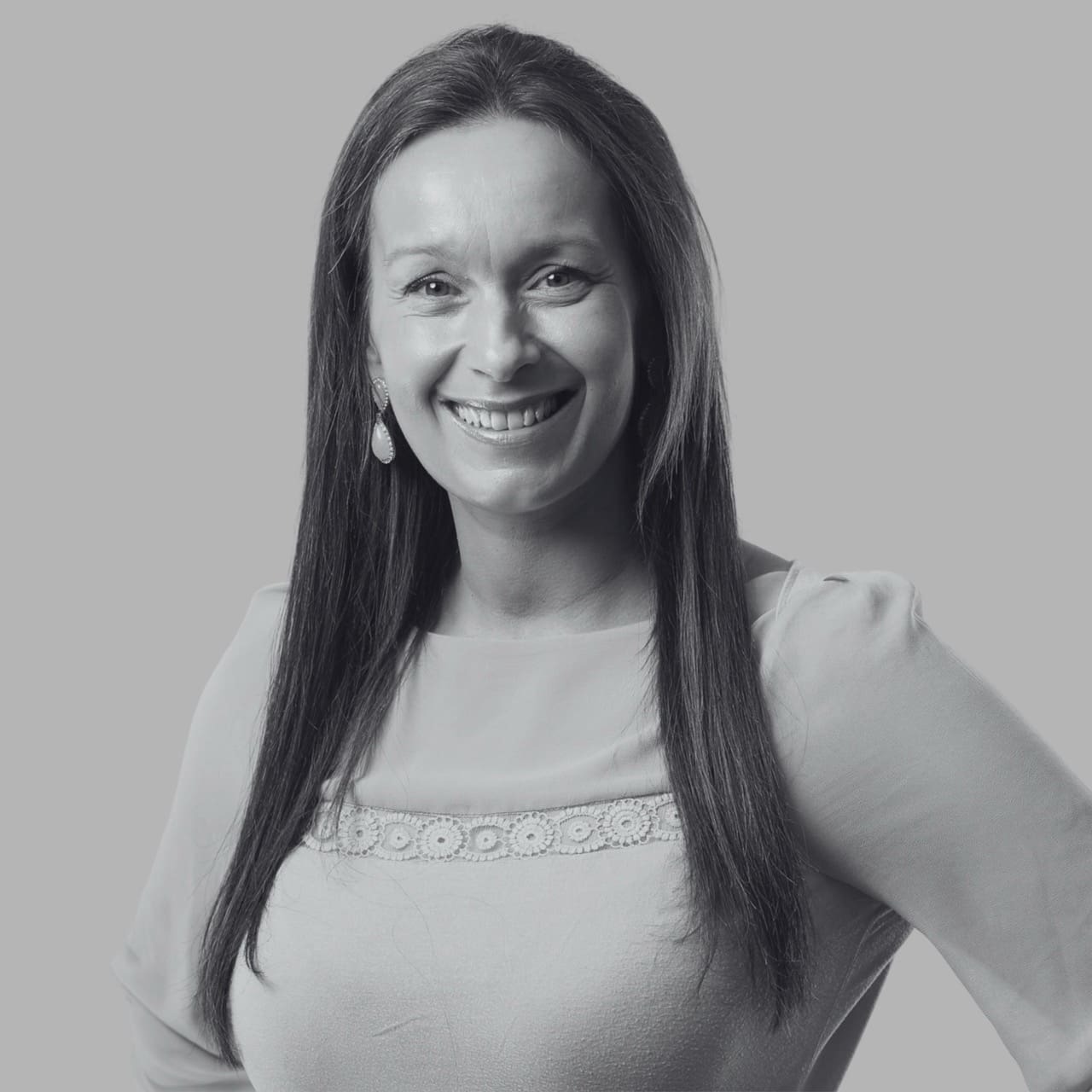 A smiling woman with long hair, wearing a light-colored top and earrings, against a grey background.