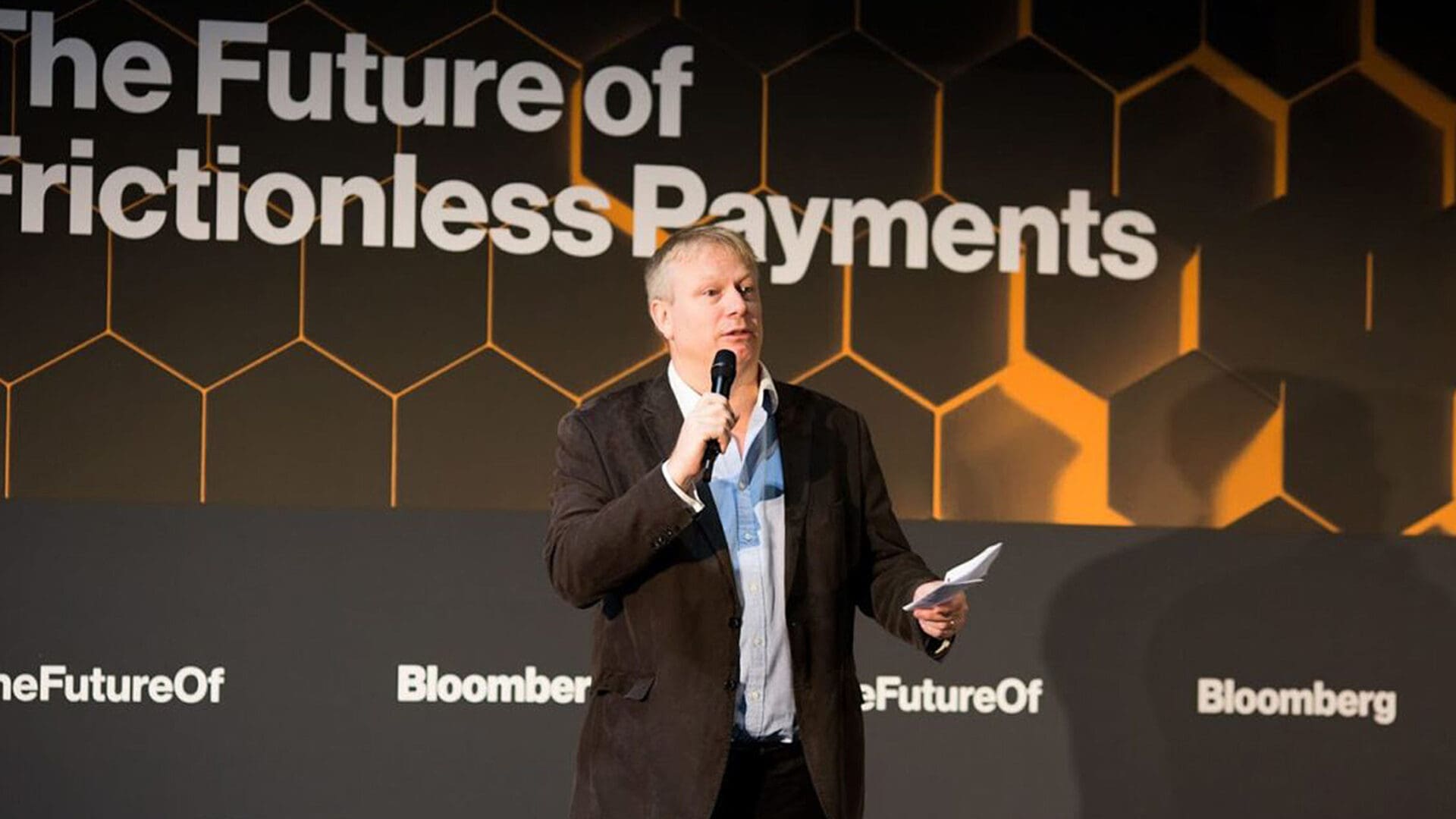 A presenter stands on stage speaking into a microphone, holding papers in one hand. Behind him, a backdrop features the text "The Future of Frictionless Payments" along with the Bloomberg logo and the hashtag "#TheFutureOf." The background design includes