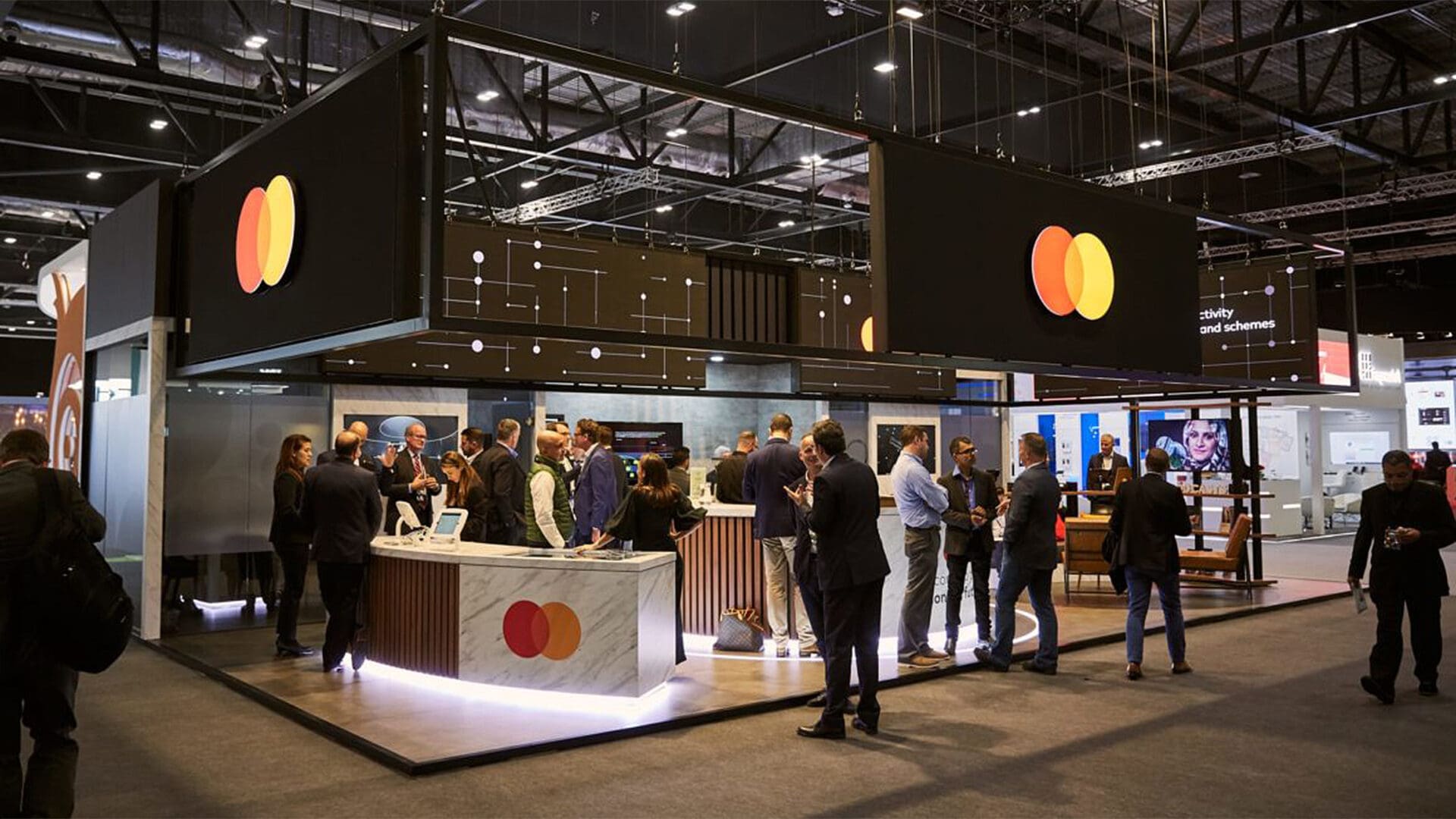 A busy exhibition area featuring a prominent Mastercard booth with a large logo display. Several attendees are engaged in conversations around a central booth with a marble countertop, while other booths and displays are visible in the background.