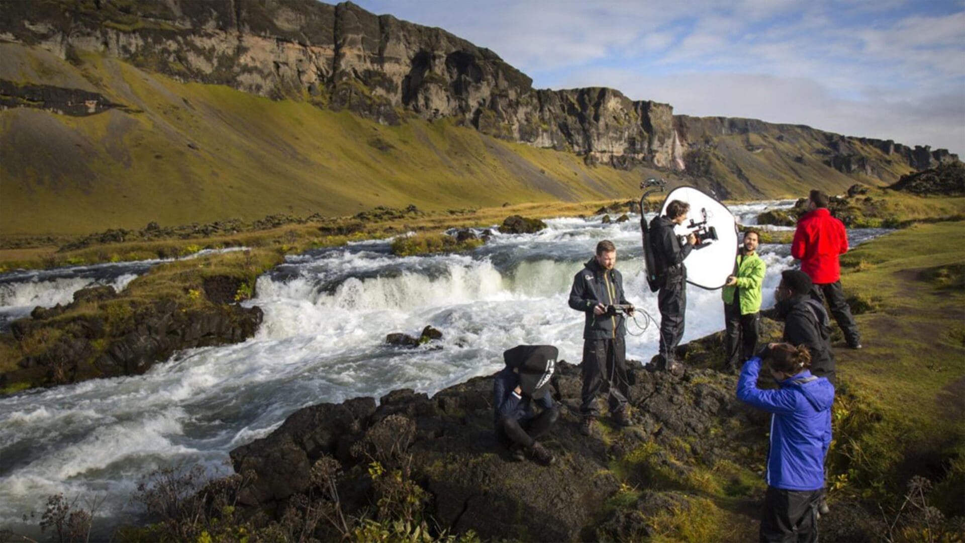 A film crew works by a rushing river in a scenic landscape, using lighting and video equipment.