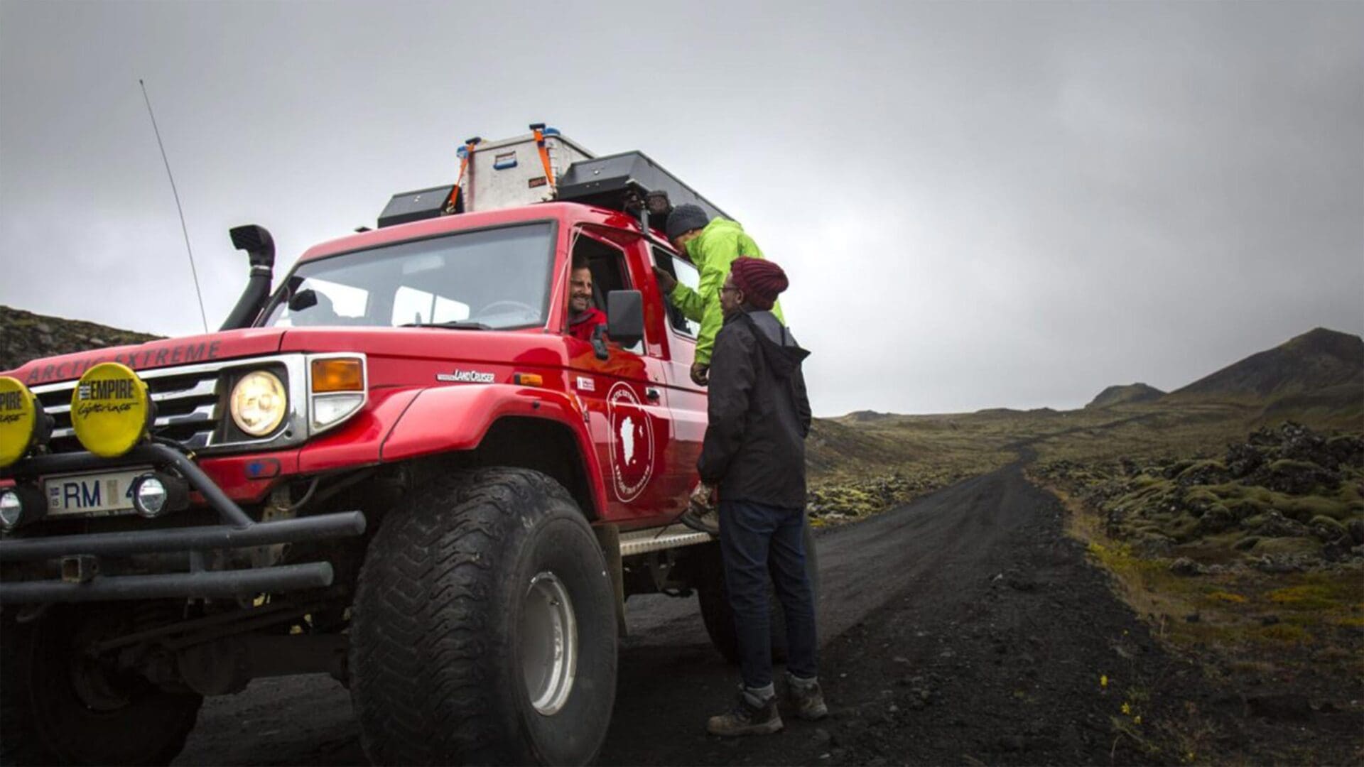 A red Toyota Land Cruiser, labeled "ARCH EXTREME" and "Empire Experience," is parked on a rocky road in a mountainous landscape. Two people are interacting by the vehicle; one is leaning into the driver's window, while the other stands beside